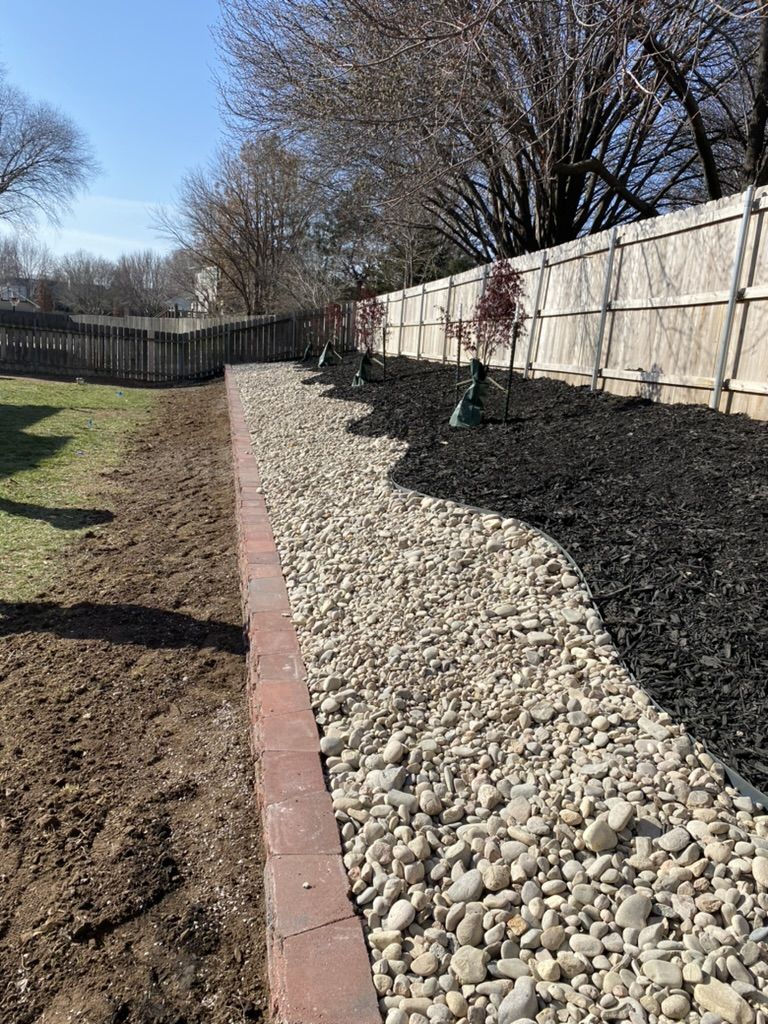 Retaining wall running along the fence with a curved river rock border and black mulch