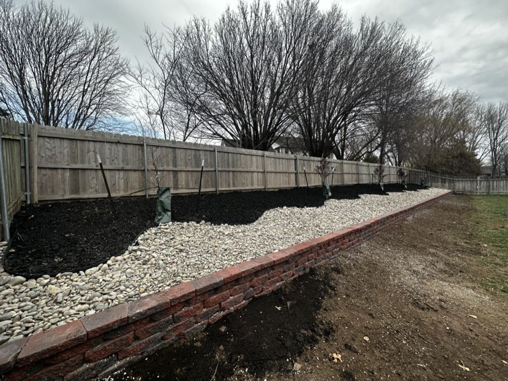 Long block retaining wall in the back yard with river rock, black mulch and young trees along the fence